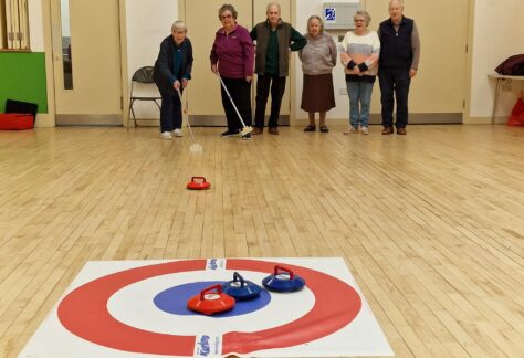 A group of people standing behind a mat marked with circles. One of them is pushing a kurling stone towards the mat.