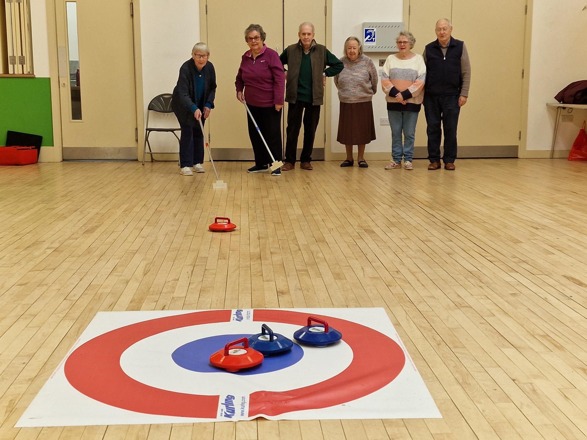 A group of people standing behind a mat marked with circles. One of them is pushing a kurling stone towards the mat.